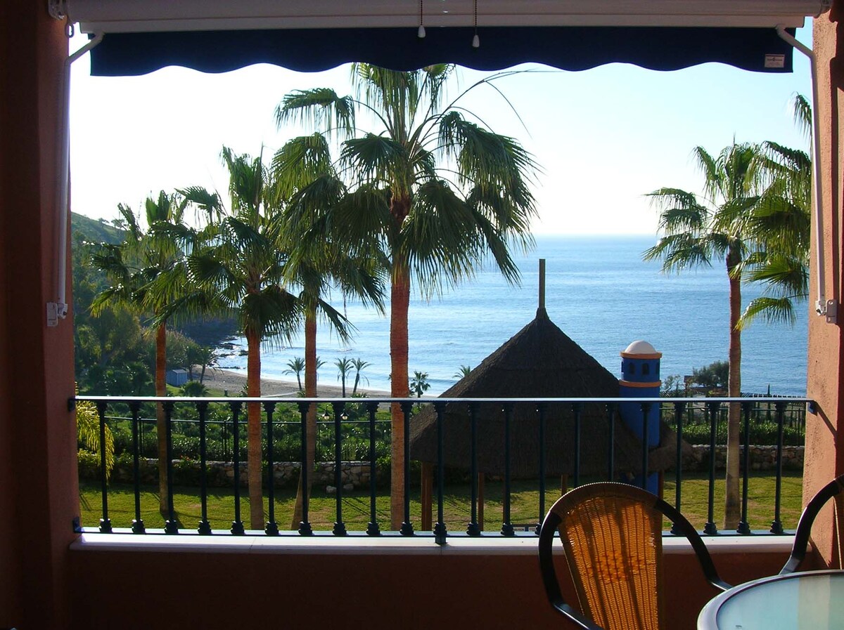 A view from a terrace showcases a serene landscape with palm trees and a clear view of the sea. The horizon blends into the sky, while a thatched-roof structure and a blue tower are visible in the foreground, framed by a decorative railing.