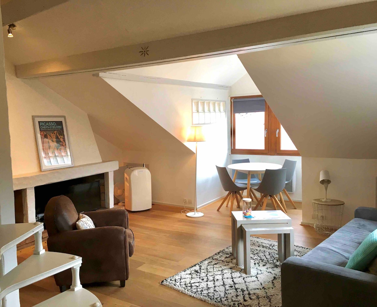A spacious living area features a comfortable seating arrangement with a dark armchair and a grey sofa, complemented by a light wooden coffee table. A round dining table with grey chairs is positioned by windows, allowing natural light to fill the space.