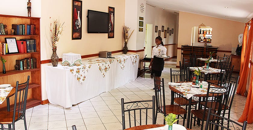 The dining area features a series of neatly arranged round tables, each adorned with tablecloths and centerpieces. A buffet table is set against one wall, complemented by a bookshelf and a television. A staff member is visible, assisting in the space.
