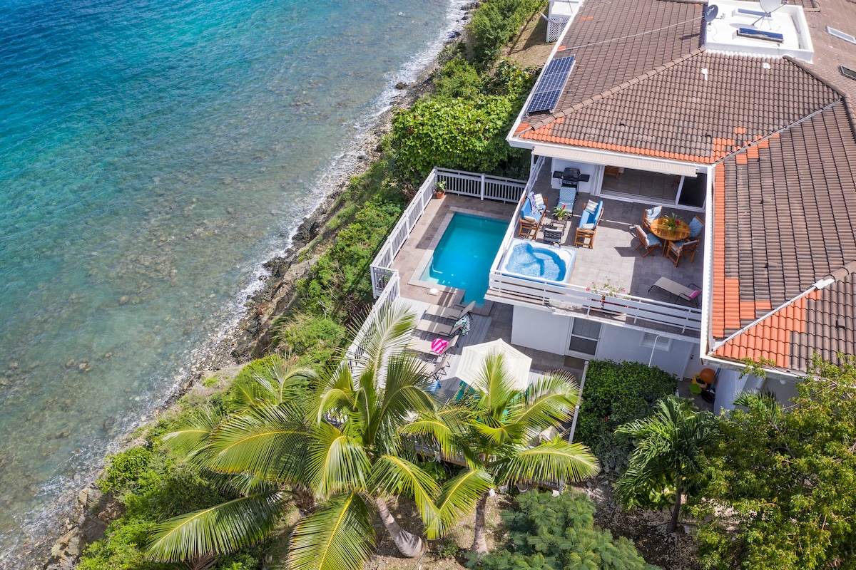 An aerial view showcases the villa positioned along the coastline, featuring a private pool and hot tub on the spacious deck. Lush greenery and palm trees surround the property, with the crystal-clear water visible just beyond the rocky shore.