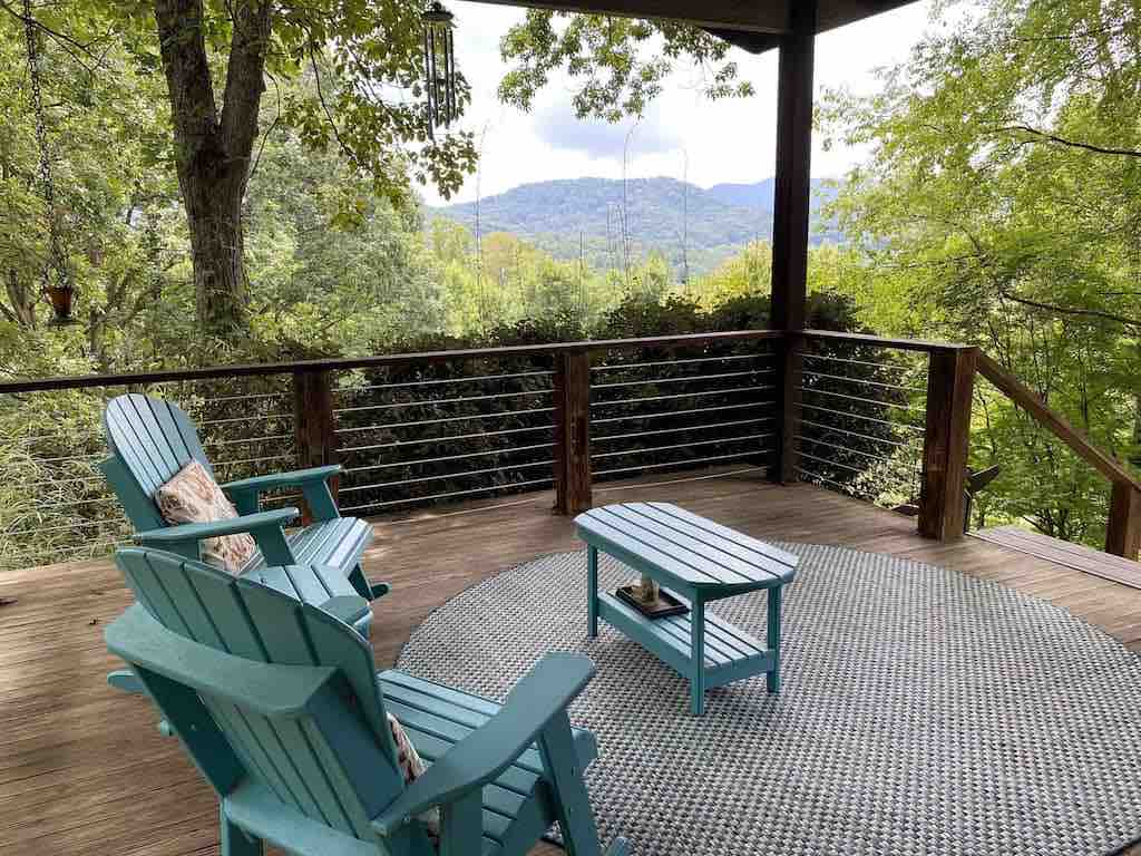 A spacious outdoor deck is furnished with two blue Adirondack chairs and a small coffee table. A woven outdoor rug lies beneath, and a natural backdrop of trees and distant mountains is visible, providing a serene setting for relaxation.