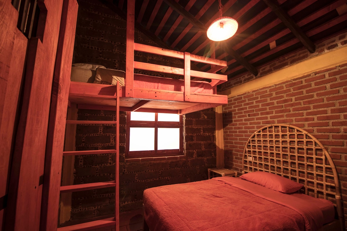A cozy sleeping area is characterized by warm red tones. A spacious bed occupies one side, while a lofted bed is positioned above, accessible by a wooden ladder. Exposed brick walls complement the rustic ceiling beams, and a window allows natural light to enter.