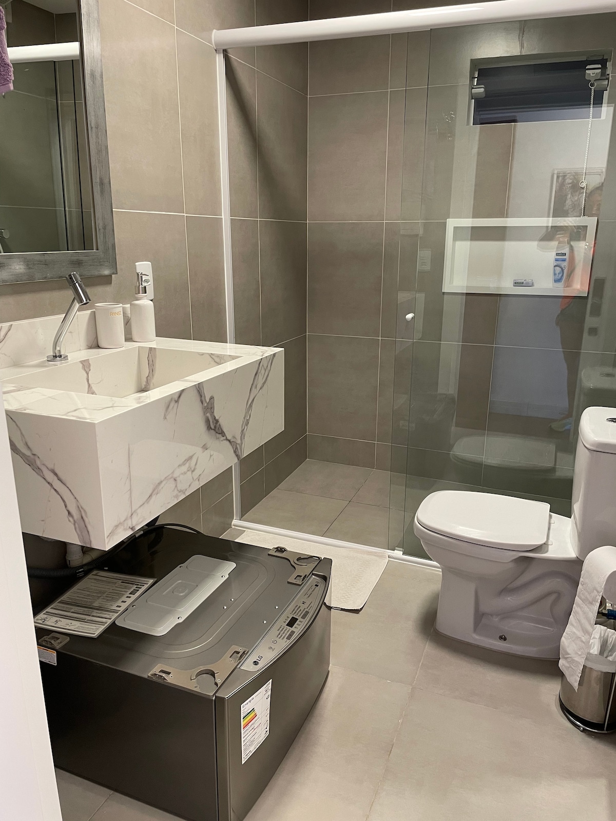 The bathroom features a modern design with a sleek countertop sink and large mirror. A glass-enclosed shower is visible alongside a washing machine placed under the sink area. Neutral tiles cover the walls and floor, creating a clean and functional space.