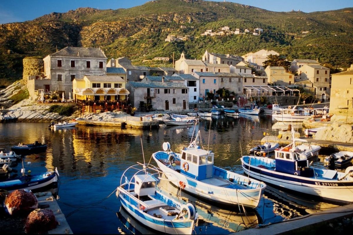 A serene harbor scene captures traditional fishing boats docked beside quaint stone buildings. The reflections on the calm water showcase the surrounding hills, while the warm sunlight bathes the village in a gentle glow.