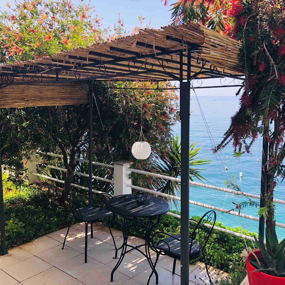 An outdoor seating area is showcased with a small black table and two chairs, surrounded by lush greenery. A wooden pergola provides shade, while a hanging lantern adds a subtle lighting element. The sea is visible in the background, enhancing the tranquil atmosphere.