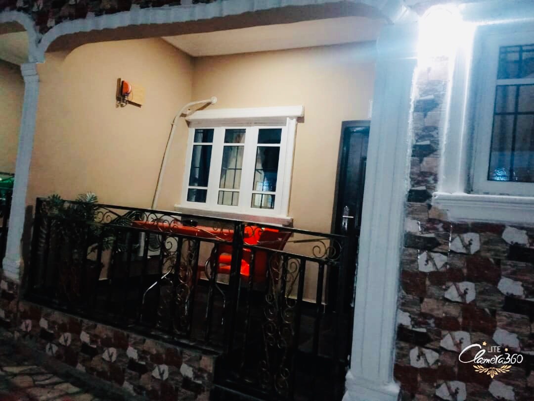 A covered porch area features a small table and two red chairs, providing a comfortable seating space. Decorative stonework frames the entrance, while a light fixture adds illumination. A view of the window showcases the interior subtly beyond.