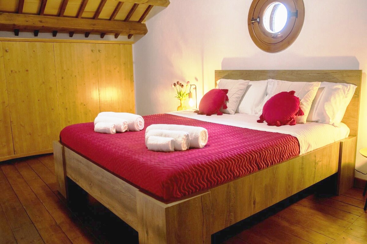 A cozy sleeping area features a large bed with a red patterned coverlet and decorative pillows in shades of red. Soft, warm lighting enhances the wood accents, while a round window allows natural light to enter. Wooden cabinetry lines the wall, contributing to the inviting atmosphere.