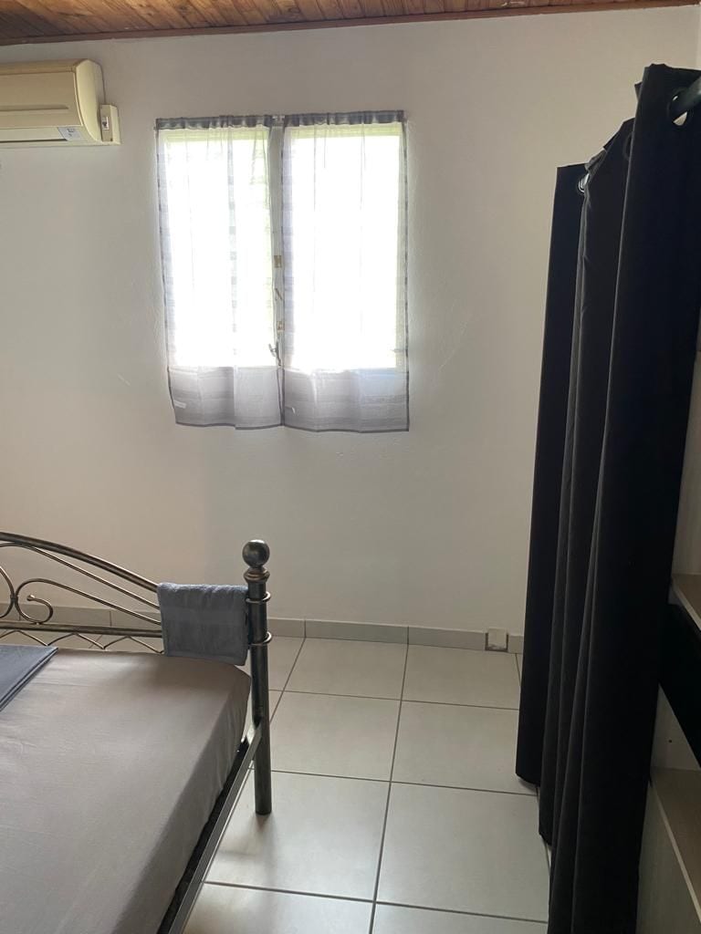 A light-filled corner of a bedroom features a modest metal bed with a soft mattress. Two windows with sheer curtains allow natural light to brighten the space, complemented by a small, dark privacy curtain.