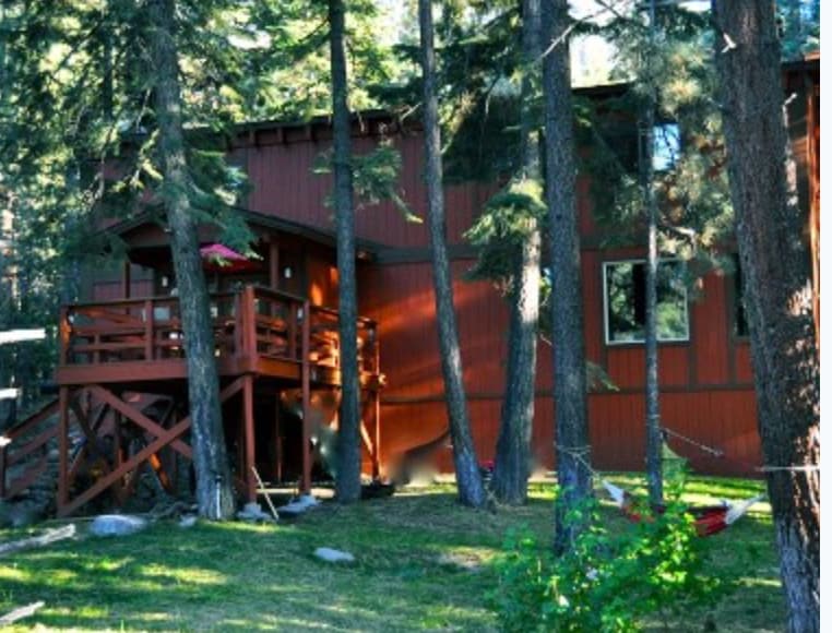 A rustic cabin with red wooden siding is nestled among tall trees. A spacious deck with a railing offers outdoor seating. The surrounding grassy area features large rocks and showcases a peaceful forest environment, creating a serene retreat.