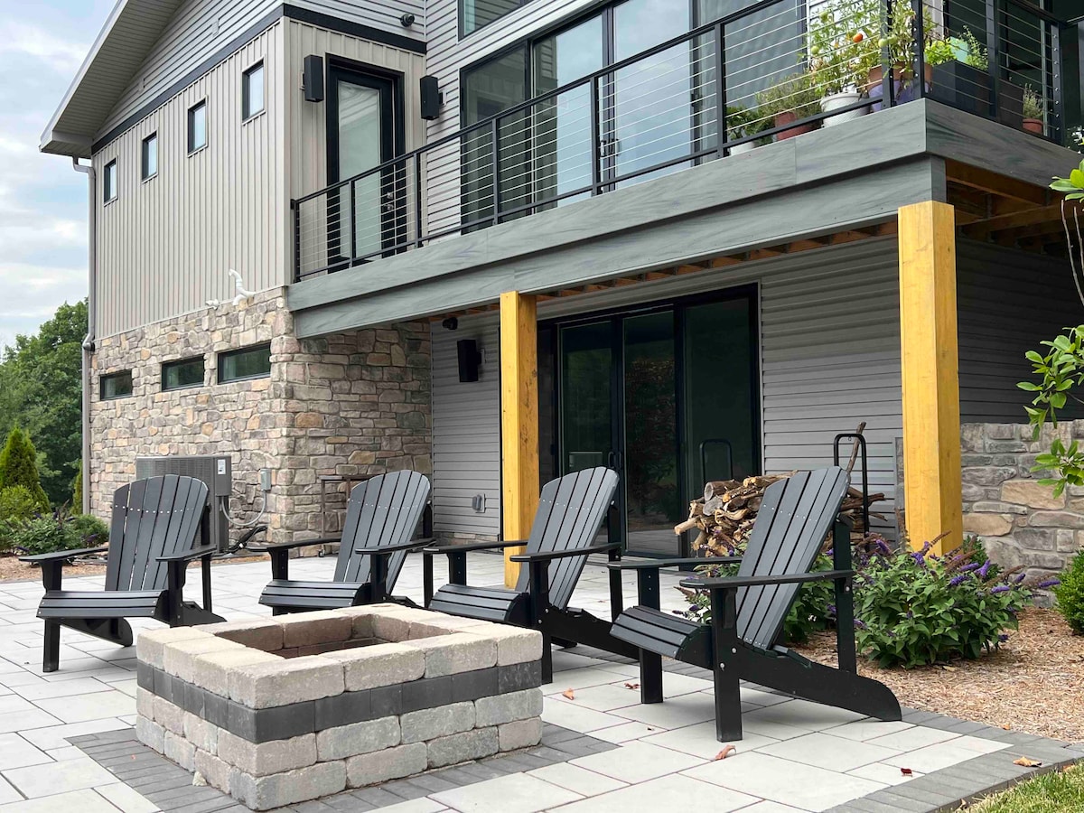 An outdoor seating area features four black Adirondack chairs arranged around a stone fire pit. The modern home in the background showcases large glass sliding doors and wooden support beams. Planters with greenery add a touch of nature to the contemporary design.