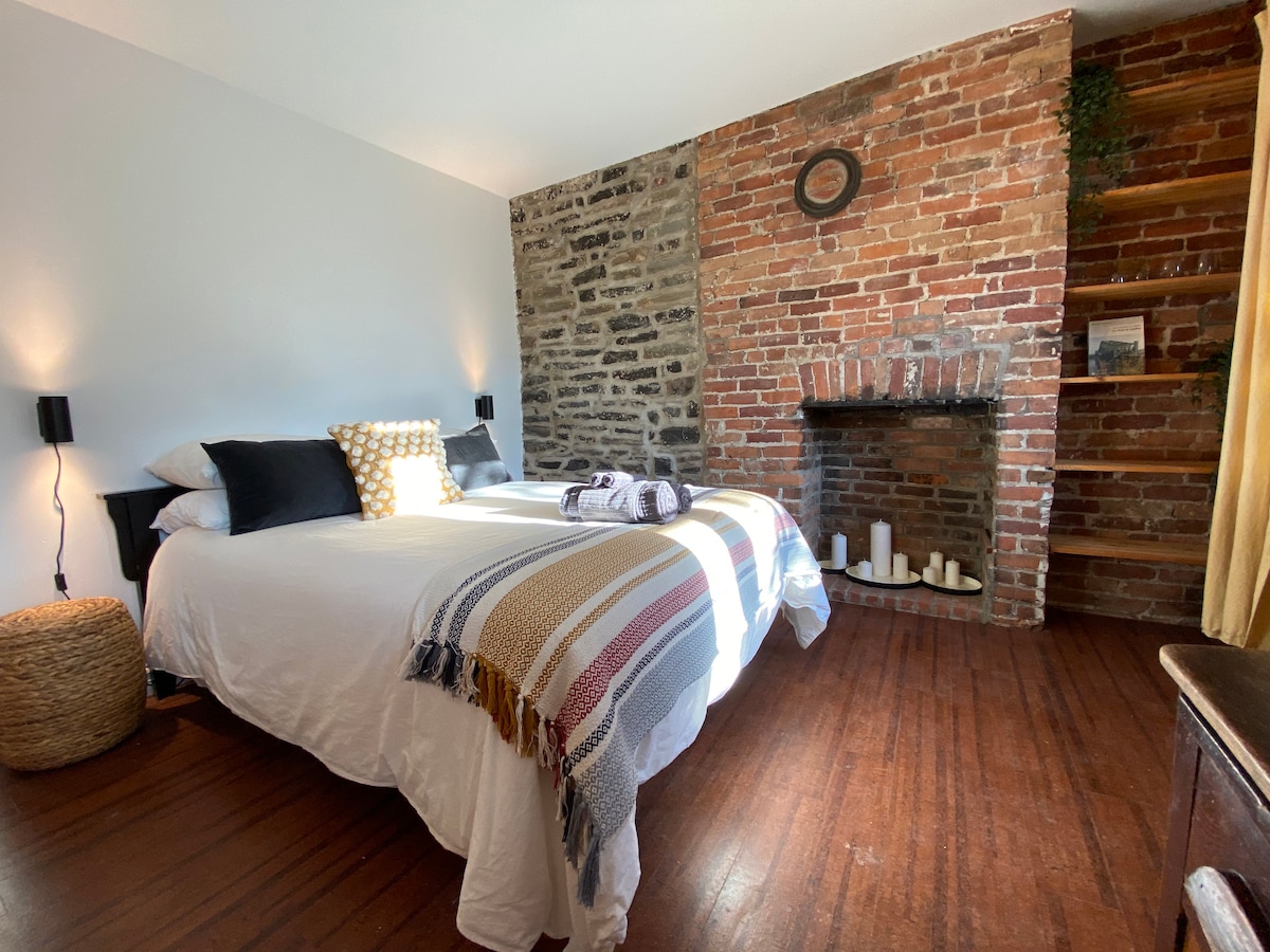 A spacious sleeping area features a comfortable bed adorned with decorative pillows. A textured wall of exposed brick and stone creates a cozy ambiance. Decorative candles are arranged thoughtfully on the fireplace mantle, enhancing the charm of the room.