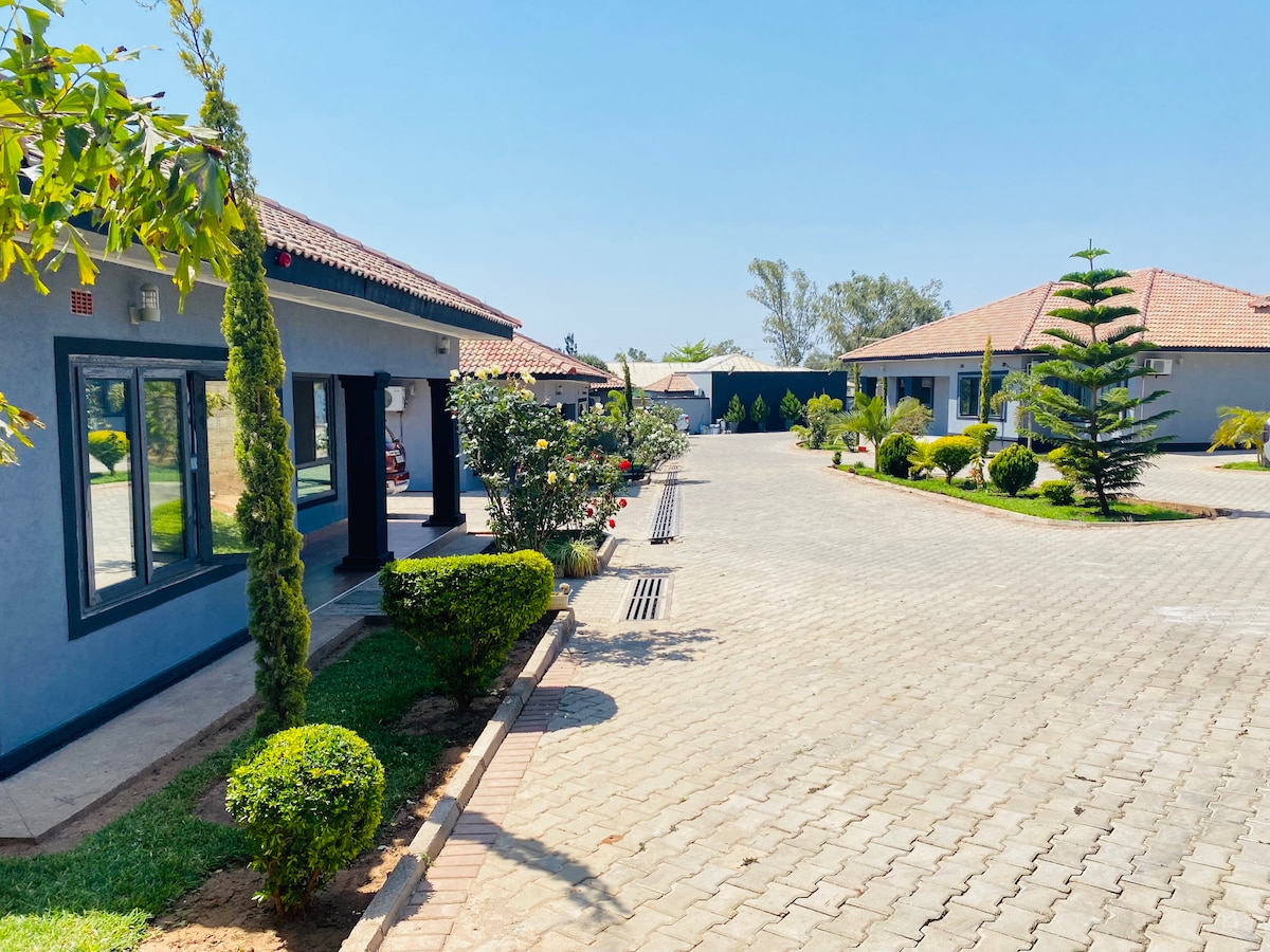 The image displays a serene courtyard featuring well-maintained landscaping with neatly trimmed bushes and trees. Two apartment buildings are visible, each with a terracotta roof. A smooth cobblestone pathway runs through the center, guiding toward further greenery and blue sky.