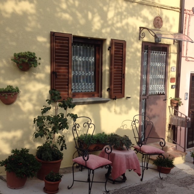 A welcoming entrance features a light yellow facade with wooden shutters on either side of a door. Two stylish chairs are positioned around a small table adorned with a pink tablecloth. Flower pots with greenery accentuate the space, creating a cozy outdoor seating area.