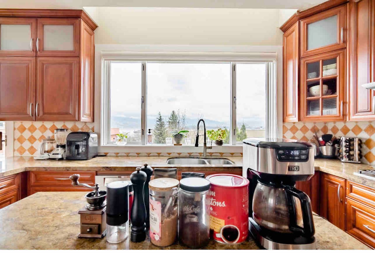 The kitchen features wooden cabinetry and a spacious countertop, with a coffee maker and a variety of canisters organized neatly. A large window offers natural light and provides a view of the outdoors. Essential appliances are also visible, enhancing the functionality of the space.