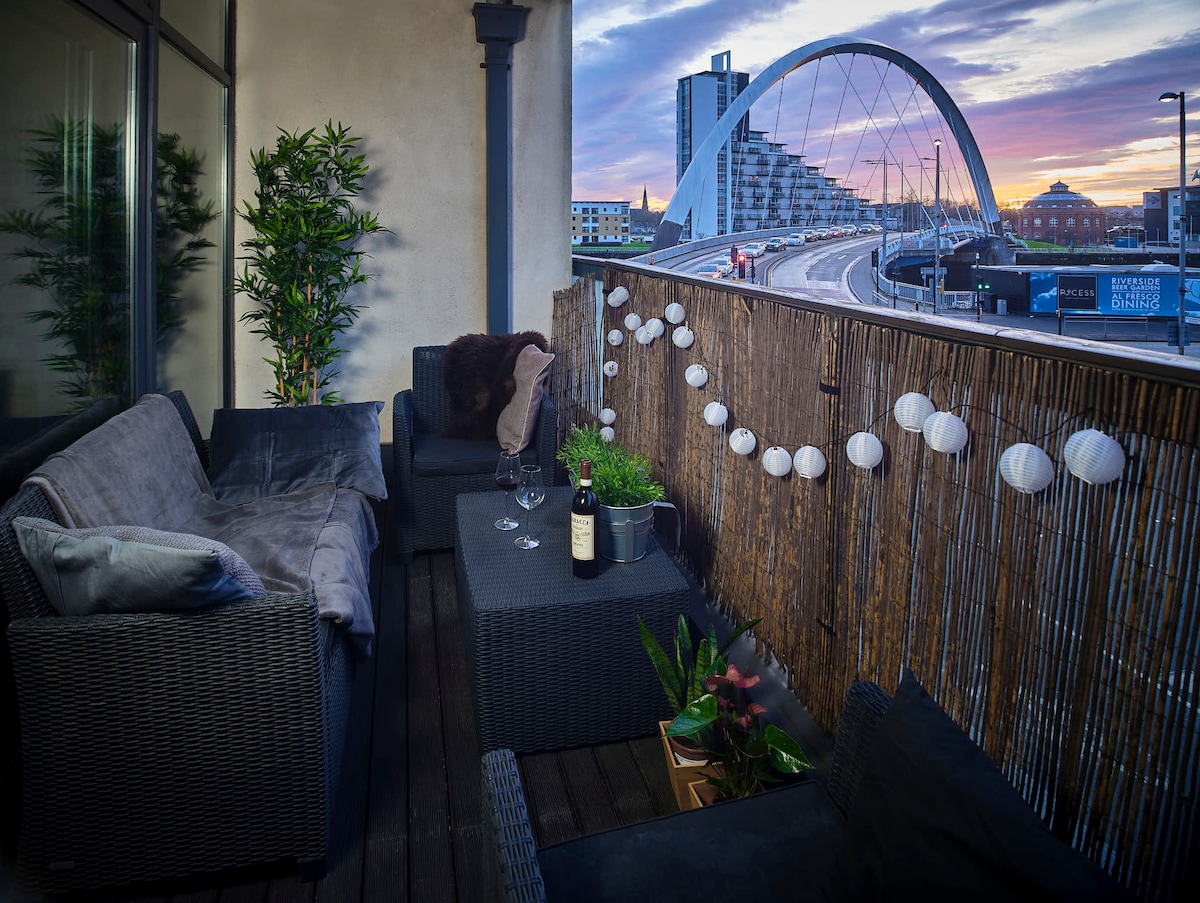 A spacious balcony features outdoor seating surrounded by greenery and decorative string lights. A coffee table with a bottle of wine and glasses is positioned in the center, providing a cozy setting to enjoy views of the Clyde Arc and a vibrant sunset.