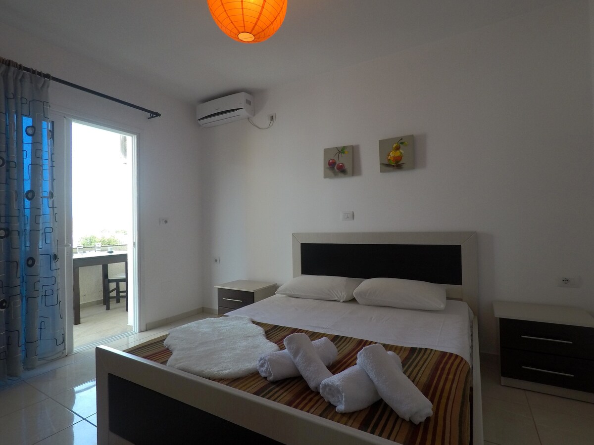 A bright bedroom features a large bed with white linens and a striped throw. Decorative wall art depicting birds is positioned above the bed. A doorway leads to a small balcony, with sheer blue curtains allowing light to enter the space.