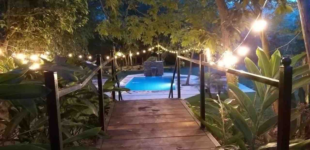 A wooden bridge leads to an inviting pool area, illuminated by string lights hanging from surrounding trees. Lush greenery frames the scene, offering a peaceful ambiance. The pool, tucked behind the foliage, reflects soft lights, enhancing the outdoor setting.
