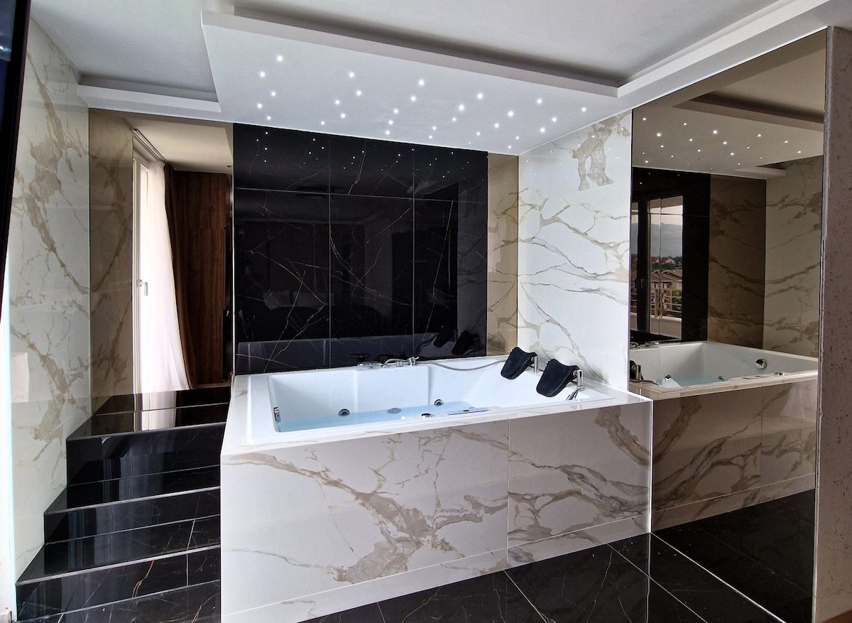 An elegant bathroom features a modern bathtub surrounded by marble walls with stylish accents. Soft, ambient lighting is provided by recessed ceiling lights. A large mirror enhances the space, reflecting the luxurious design elements and creating a sense of openness.