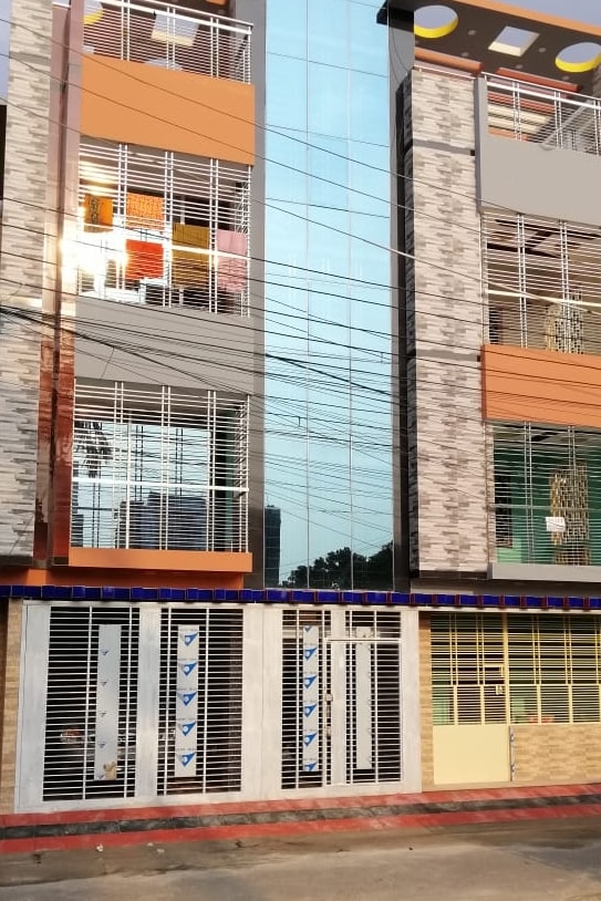 A modern three-story building features a sleek glass facade with horizontal lines and stone accents. The entrance is framed by decorative metal grilles, while multiple windows allow for natural light. The exterior is complemented by colorful furnishings visible through the upper windows.
