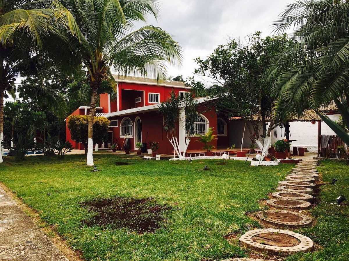 A spacious red house is set amidst a lush green garden, featuring tall palm trees and a stone pathway that leads to the front entrance. The property showcases a large outdoor area, providing ample space for relaxation.