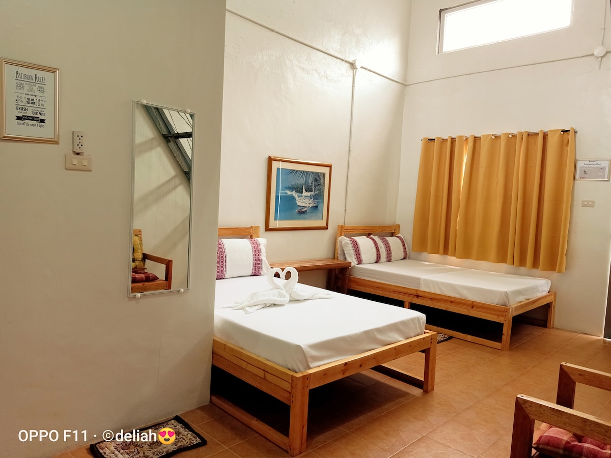 The room features two spacious beds with wooden frames and fresh linens. A mirror reflects the bright ambiance, while a piece of artwork adds a touch of decor. Yellow curtains allow natural light, complemented by an inviting floor and simple furnishings.