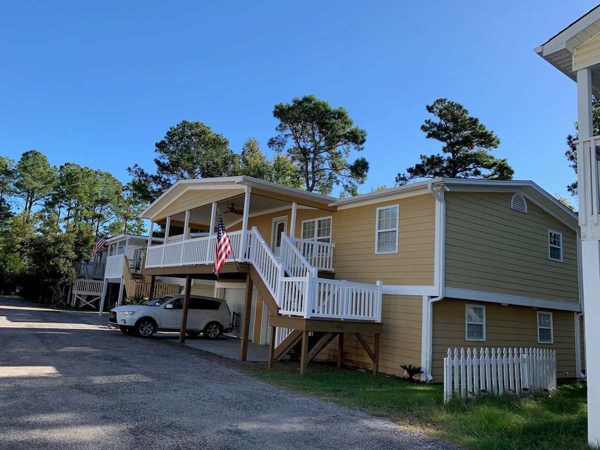 The exterior of the home displays a welcoming facade with yellow siding and white railings. Flags are seen hanging from the porch, and a gravel driveway leads to a garage. Surrounding trees offer a natural backdrop in the clear blue sky.
