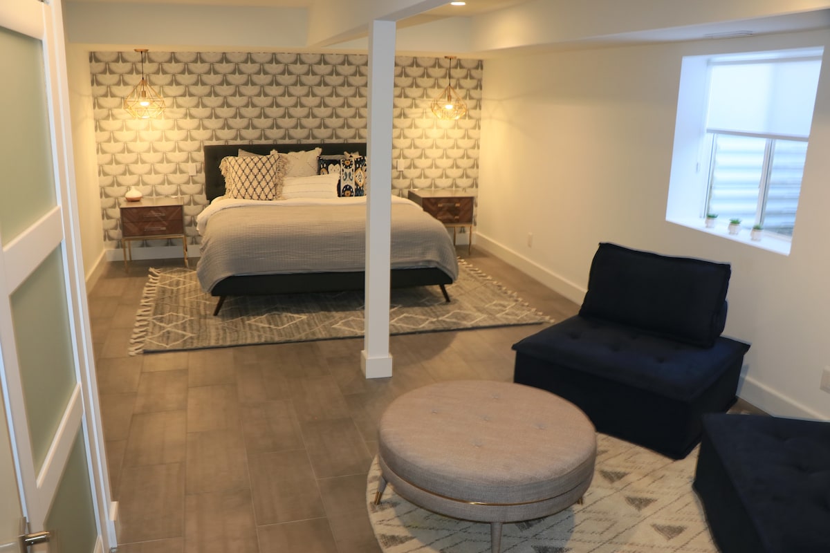 The downstairs bedroom is the largest, with its own lounge area. The accent wall features Genevieve Gorder's designer wallpaper, with two geometric, brass reading lights on each side of the luxury king mattress dressed in soft, high quality linens.