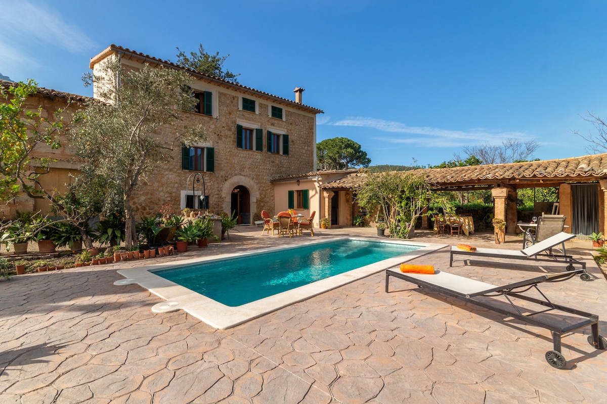 The charming holiday home features a newly constructed pool surrounded by smooth stone paving. Comfortable sun loungers are positioned beside the pool, while a shaded outdoor seating area with greenery offers a relaxing retreat. The rustic stone exterior showcases traditional architecture under a clear blue sky.