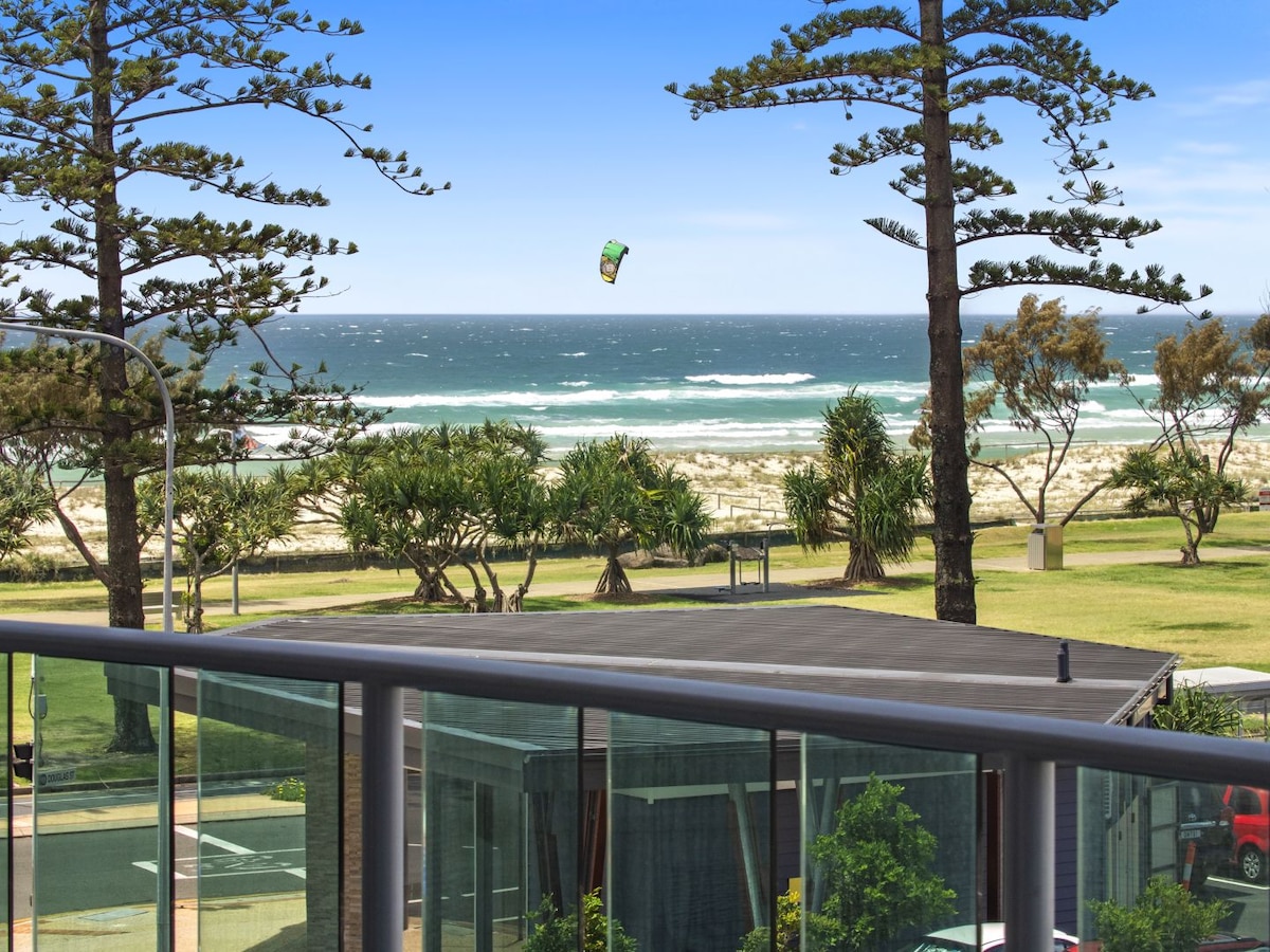 A balcony view features vibrant green palm trees and the ocean with gentle waves in the distance. A colorful kite is seen soaring above the water. The scene is framed by a modern railing that offers a sense of openness to the beachside environment.