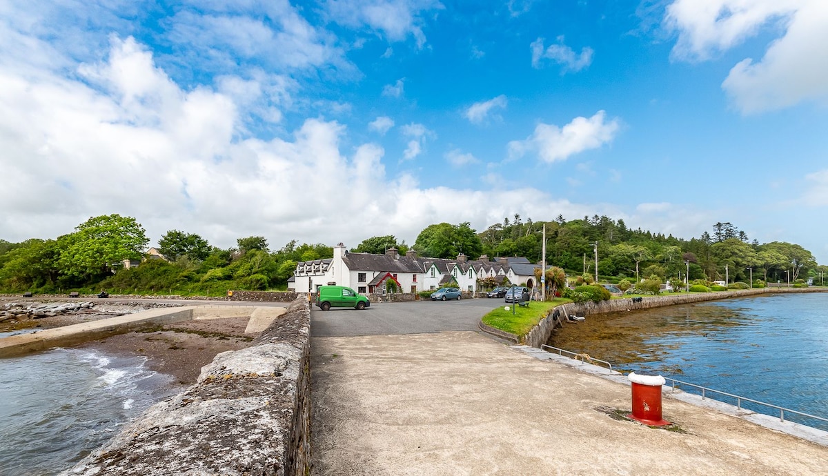 A view of the waterfront area features a concrete path leading to a cluster of traditional cottages bordered by greenery. The calm water reflects the blue sky, with a few clouds, and a parking area is visible near the cottages. A red buoy stands out near the shore.