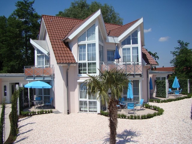 A two-story building is presented with a pink exterior and a distinctive gabled roof. Large windows allow natural light into the rooms. Outdoor seating and patio umbrellas complement the landscaped surroundings, which feature gravel pathways and palm trees.