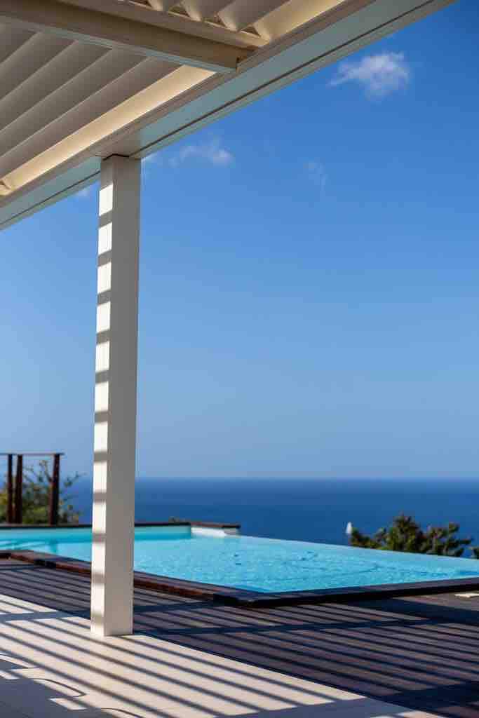 A modern infinity pool is visible under a shaded area, reflecting the blue sky and ocean. Clean lines and a wooden deck create a seamless transition from the indoor space to the tranquil outdoor setting.