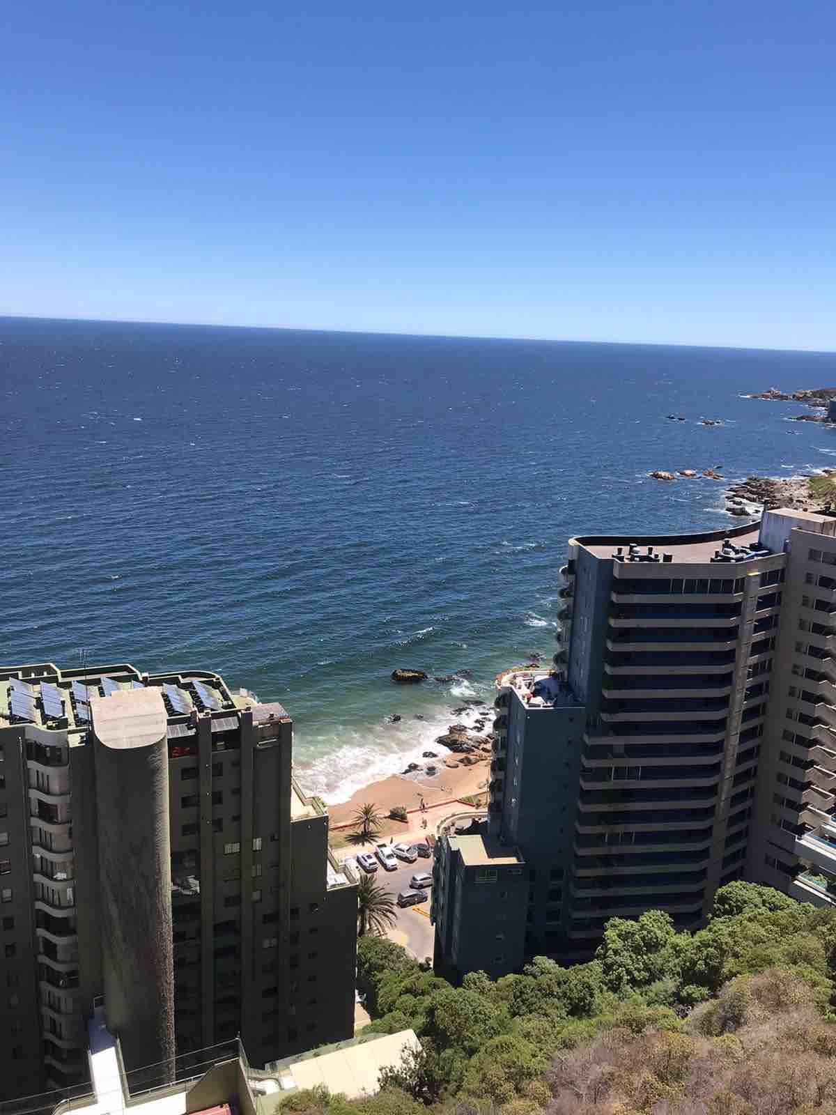 A panoramic view reveals the ocean meeting the coastline, framed by towering buildings. The deep blue water contrasts with the sandy beach below, offering a serene perspective. Small rocky formations are visible in the water, enhancing the coastal landscape's natural beauty.
