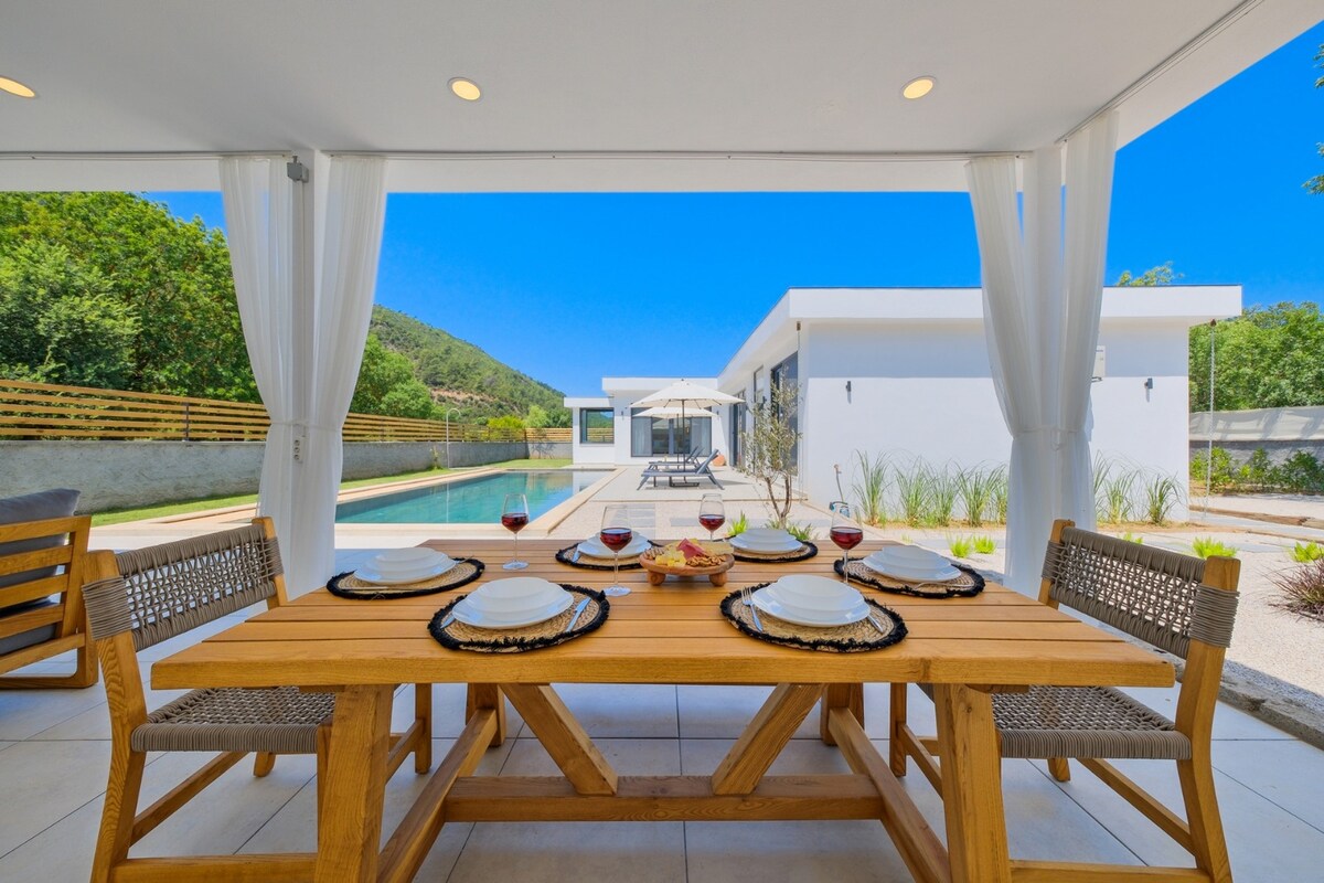 An outdoor dining area is set with a wooden table, featuring plates and glasses ready for guests. Beyond, a private pool reflects the clear blue sky, surrounded by lush greenery and a modern villa. Drapes gently frame the space, inviting relaxation.