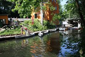 A serene waterway is depicted, bordered by lush green trees and a vibrant orange building. Several small boats are moored along the bank, alongside wooden steps leading to a pathway. People can be seen enjoying the peaceful atmosphere in this natural setting.
