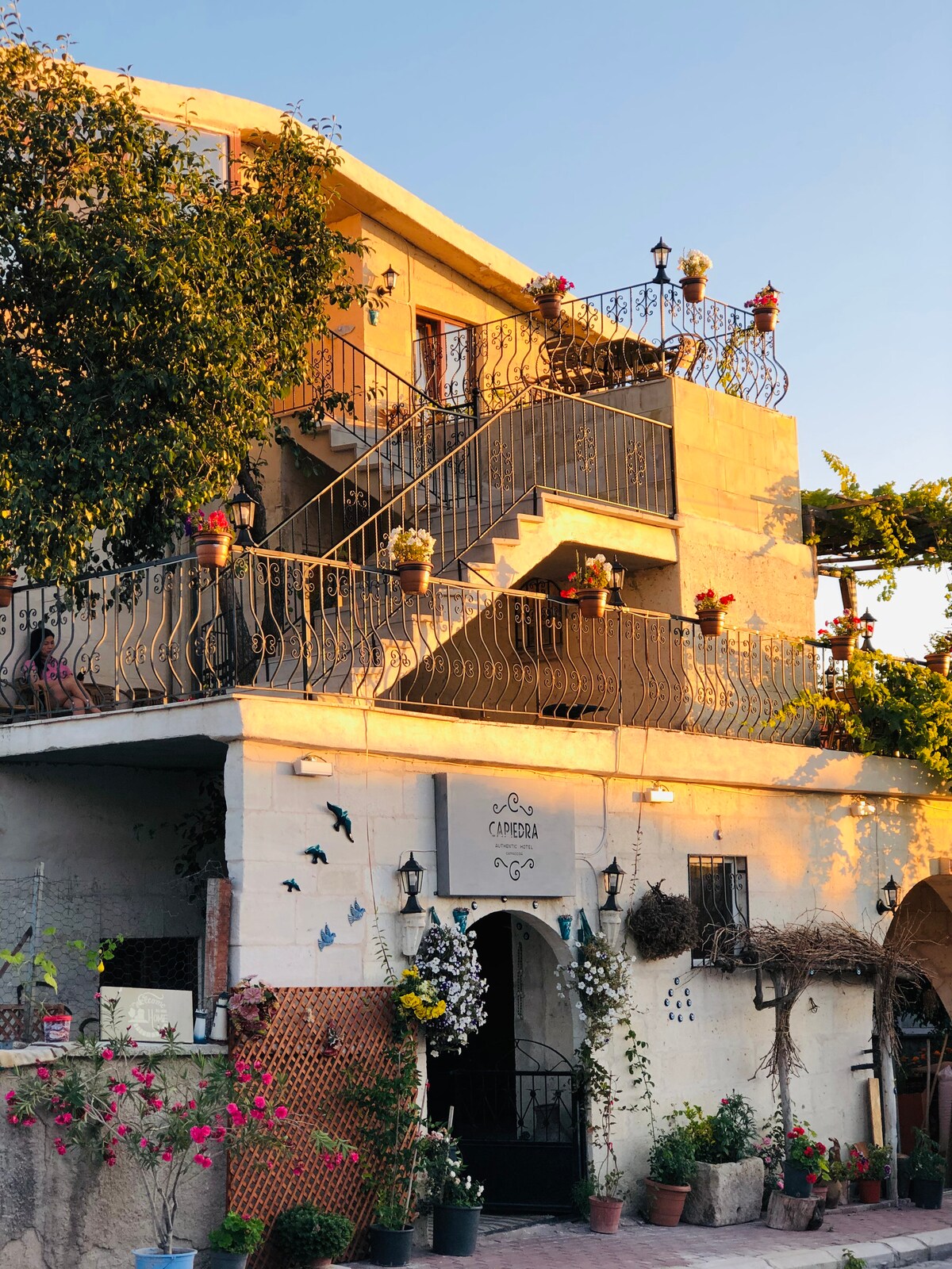The exterior of a charming multi-level building is framed by lush greenery and vibrant flowers. A decorative staircase leads to the upper level, adorned with potted plants and colorful hanging items, while the entrance showcases a welcoming sign and rustic details.