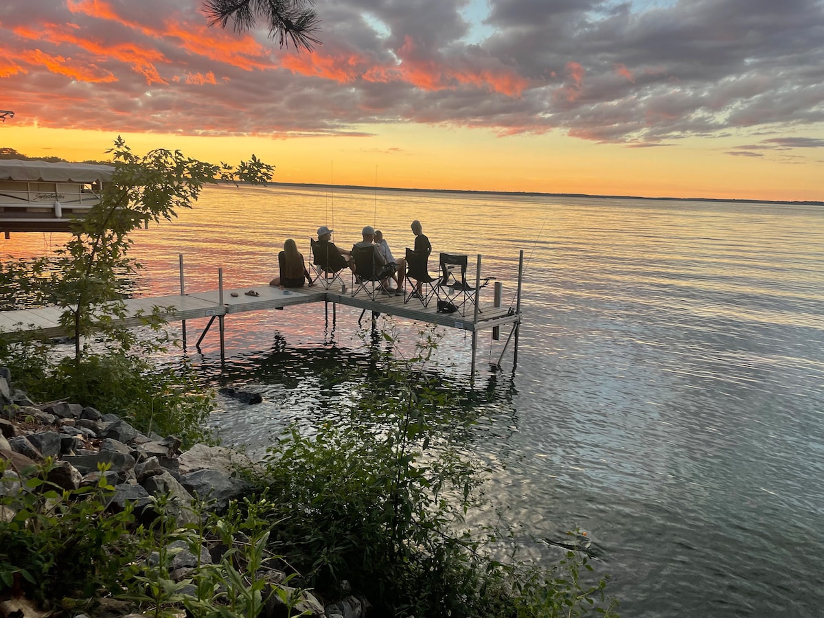 A wooden dock extends into the calm water, occupied by a group of individuals seated in folding chairs. The sunset casts vibrant hues across the sky and reflects gently on the lake's surface, surrounded by lush greenery and rocky shoreline.