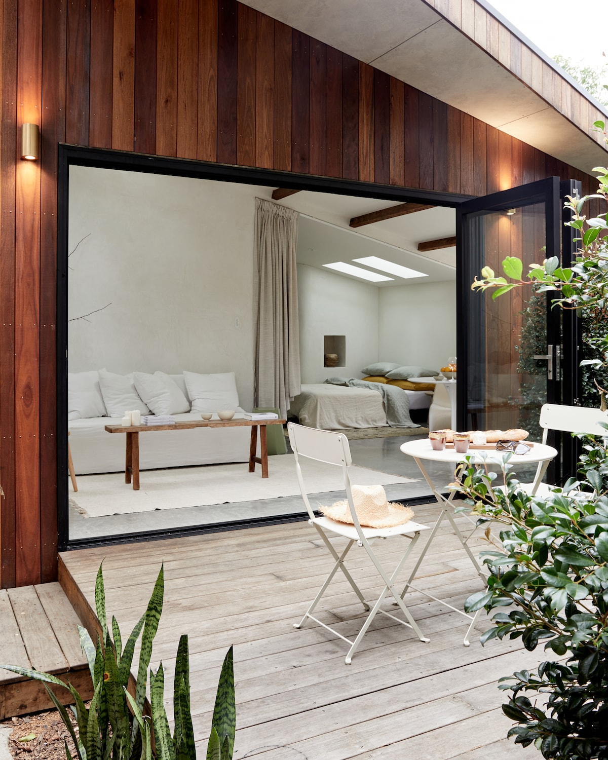 A charming outdoor seating area features a small white table with two chairs, surrounded by greenery and wooden decking. Through a large glass door, a light-filled interior is visible, highlighting a cozy living space with a sofa and a comfortable bed in soft tones.