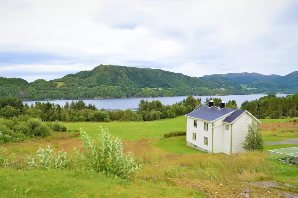 A white farmhouse is situated on a grassy landscape, overlooking a serene body of water. Lush green hills rise in the background, providing a tranquil natural setting. The sky is overcast, contributing to the calm ambiance of the scene.