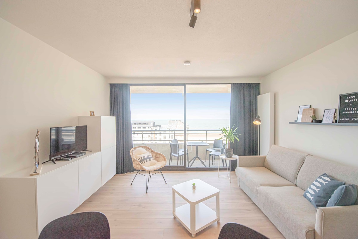 A modern living space is seen featuring a light-colored sofa and a white coffee table. A television is positioned on a media unit across from large glass doors that provide access to a private balcony. Potted plants add a touch of greenery to the inviting atmosphere.