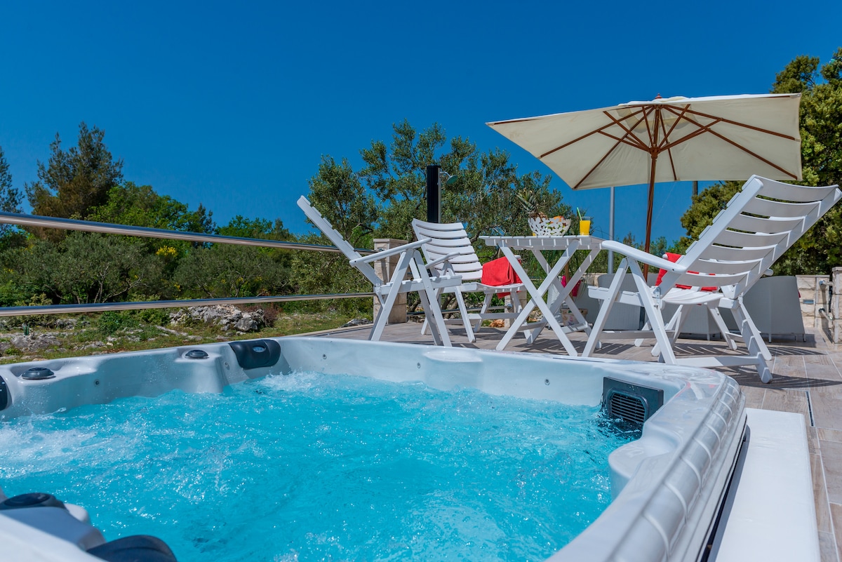 An outdoor space is showcased with a private jacuzzi filled with clear blue water and surrounded by comfortable white loungers. A large umbrella provides shade, while a dining table set with red accents is positioned nearby, framed by the lush greenery of the surrounding area.
