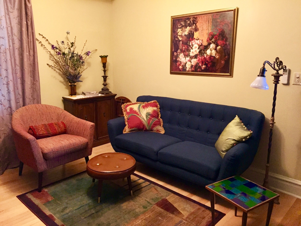 The living area features a comfortable blue sofa adorned with decorative cushions, complemented by a patterned rug. A soft pink armchair and a small round coffee table are present. A vibrant floral painting hangs on the wall, and natural light enters through the window covered by light curtains.