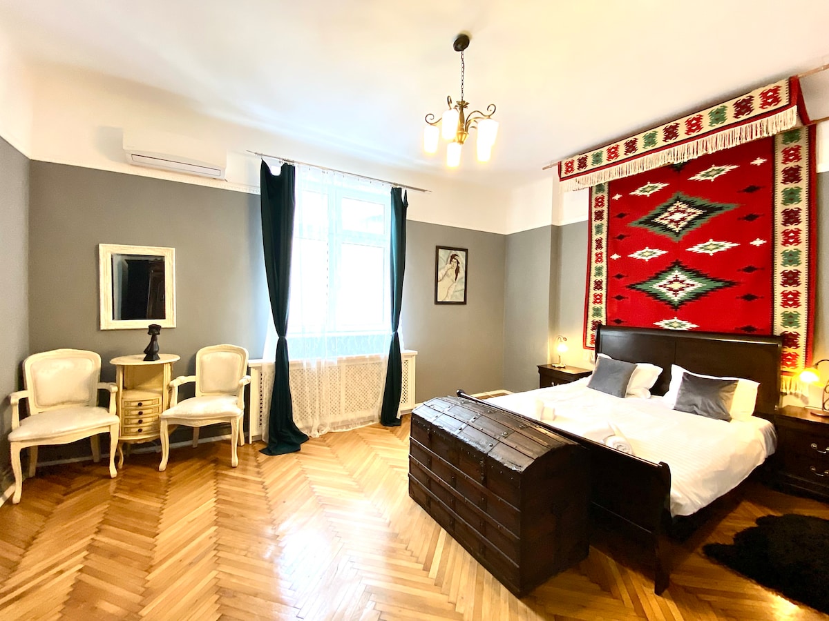 A spacious bedroom features a queen-sized bed with crisp linens, accompanied by a wooden chest at the foot. Two armchairs are positioned near a window dressed with dark curtains, while a vibrant red wall hanging adds a touch of character to the room.