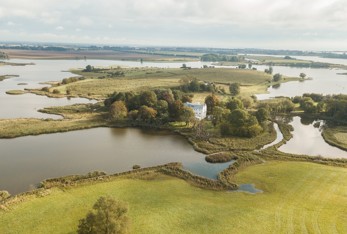 An aerial view captures the estate surrounded by tranquil waters and lush greenery. The historic house is positioned amidst a serene landscape featuring winding waterways and open fields, illustrating the natural beauty of the protected bay landscape.