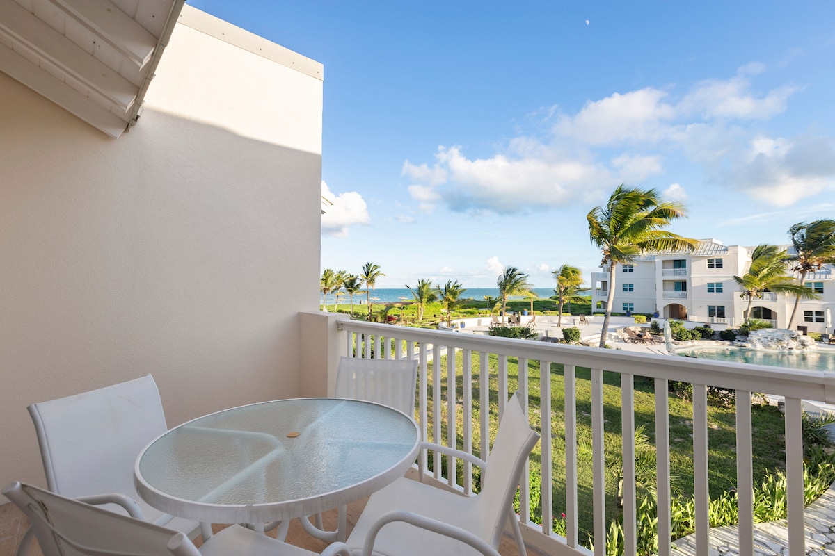 A private balcony is highlighted, featuring a round glass table surrounded by four white chairs. Scenic views of palm trees and the beach are seen in the distance, under a bright blue sky with scattered clouds.