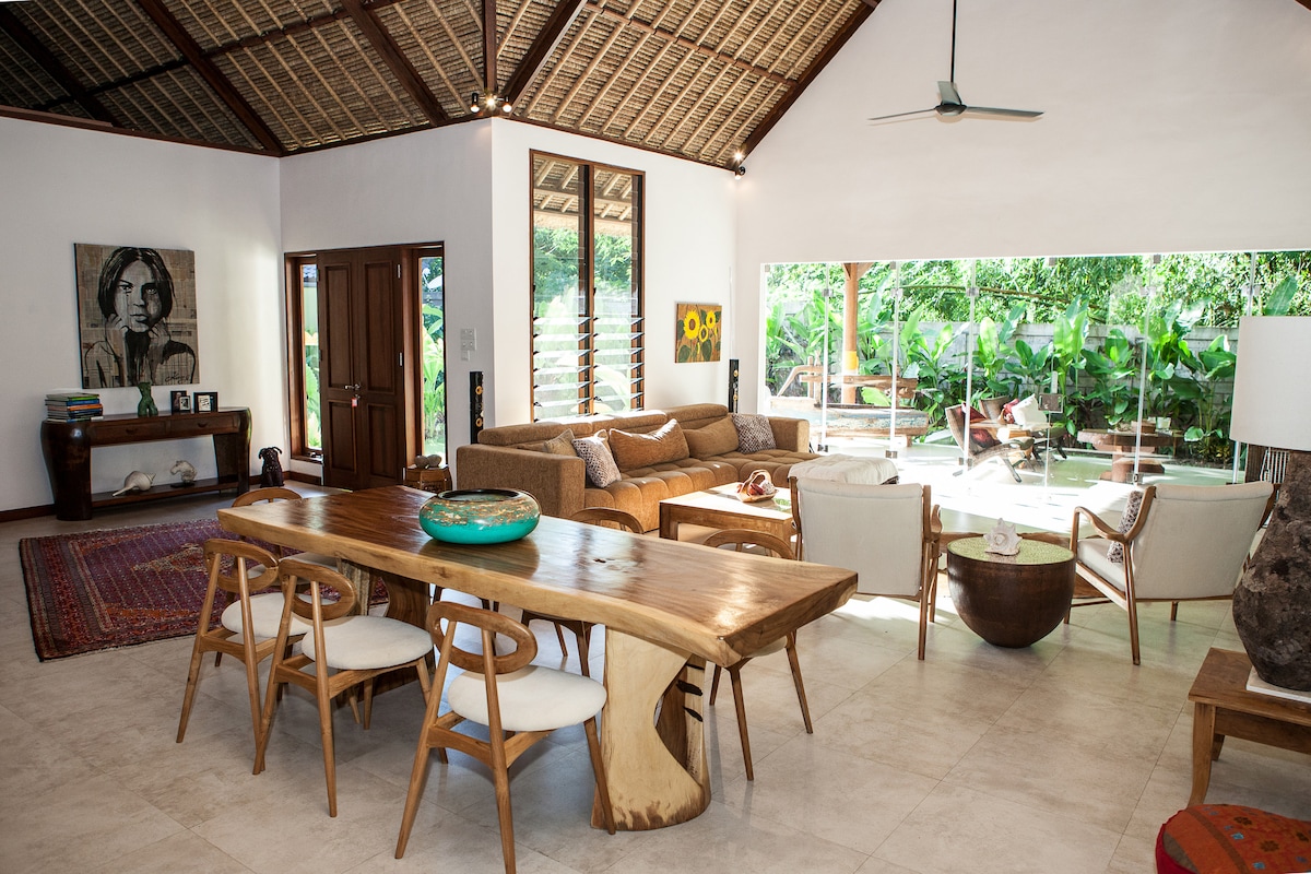 An open living space features a comfortable sofa and armchairs arranged around a coffee table. Natural light filters through large glass doors, revealing a green garden view. A rustic dining table with wooden chairs complements the warm decor, creating an inviting atmosphere.