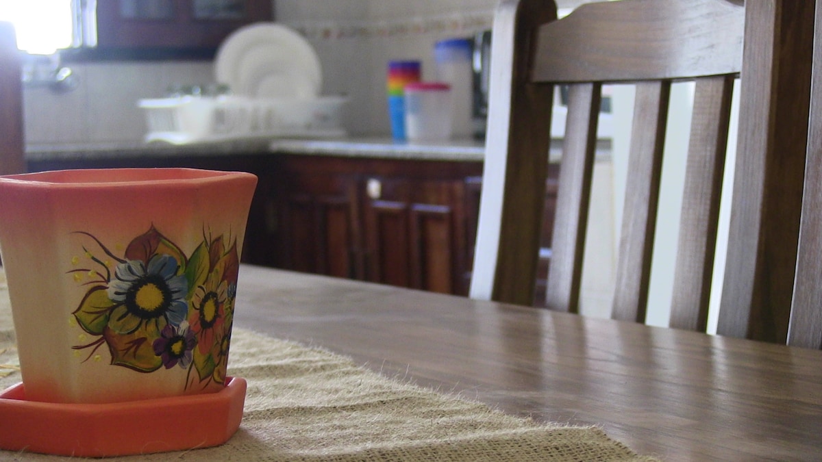 A handcrafted flower-patterned pot is positioned on a wooden table, with a woven table runner underneath. In the background, a kitchen scene is visible, featuring dishes stacked neatly by the window and a colorful kitchen item on the counter.