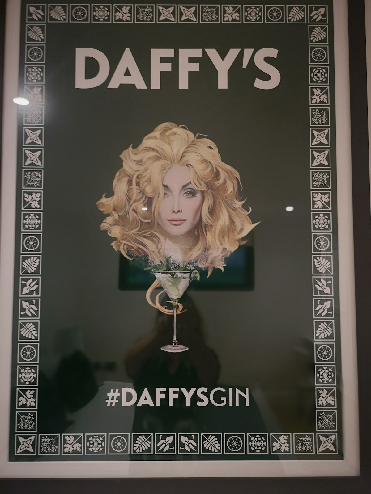 A framed poster is displayed featuring a stylized illustration of a cocktail glass topped with a garnish, accompanied by an abstract representation of a person's head with wavy hair. The predominantly green background contrasts with the white text, lending a modern touch.