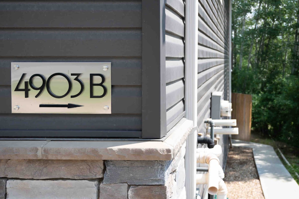 The exterior of the building is shown, featuring a clear address sign reading '4903 B.' The structure is constructed with textured gray siding and stone accents at the base. A pathway leading to the entrance is visible, bordered by greenery.