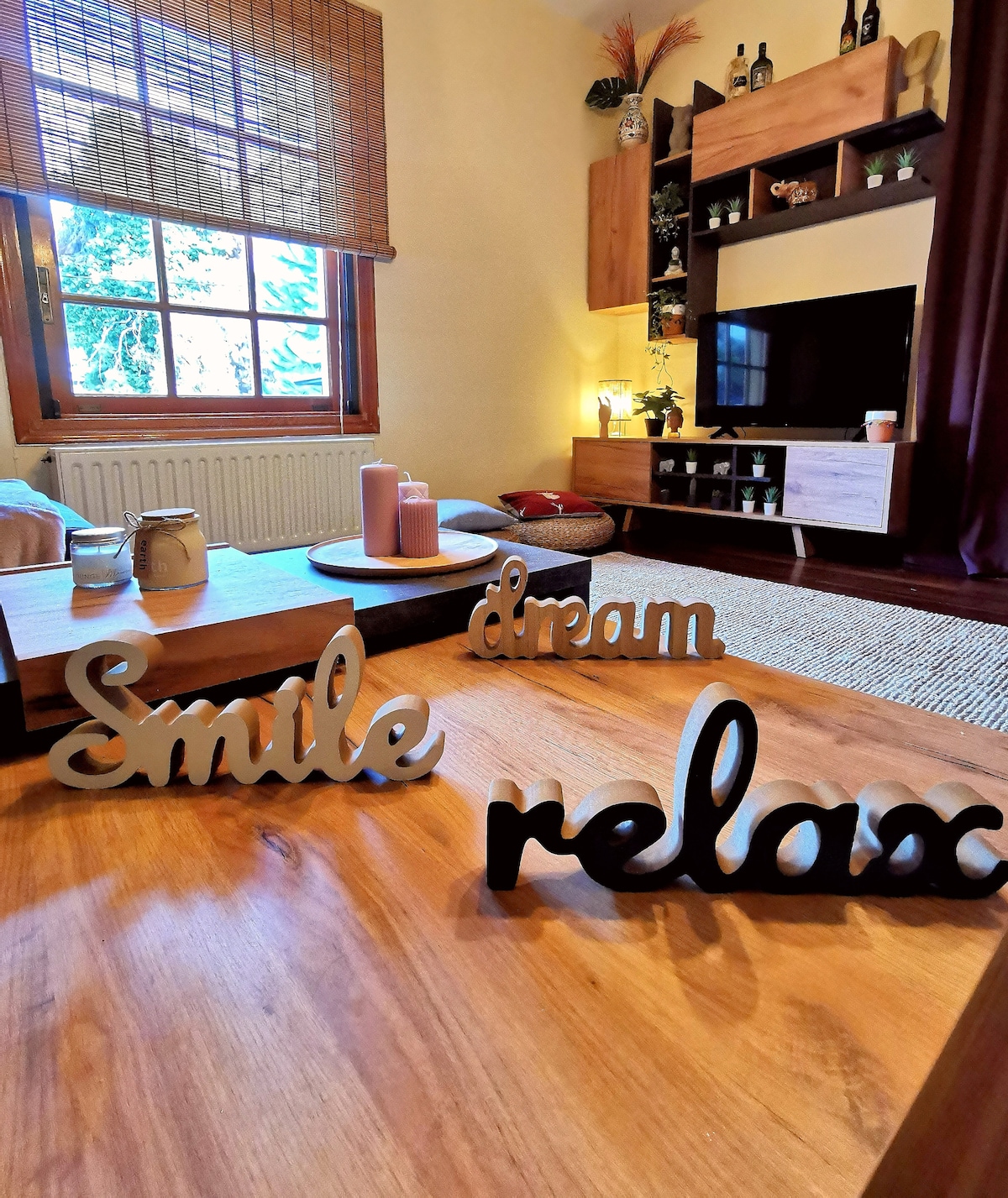 A cozy living area features a wooden coffee table with decorative text elements spelling 'Smile', 'Dream', and 'Relax'. Soft lighting creates a calming ambiance, while a television and shelves with various items are visible in the background. Natural light filters through the window adorned with bamboo shades.